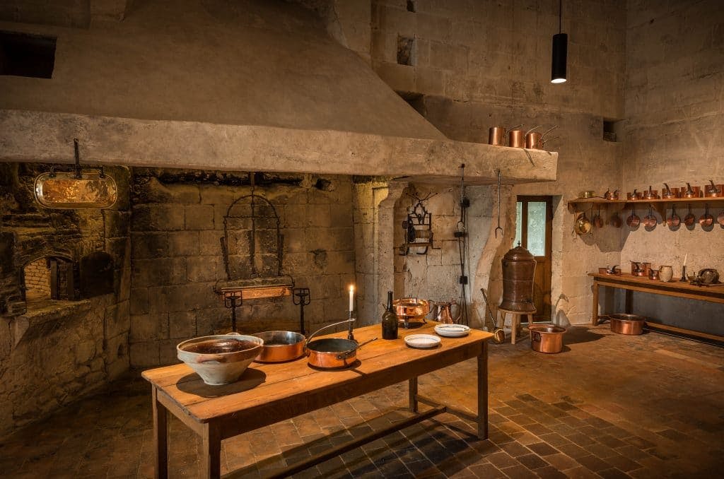 커다란 돌 벽난로, 나무 테이블, 구리 냄비, 다양한 조리도구가 있는 소박하고 고풍스러운 주방입니다.