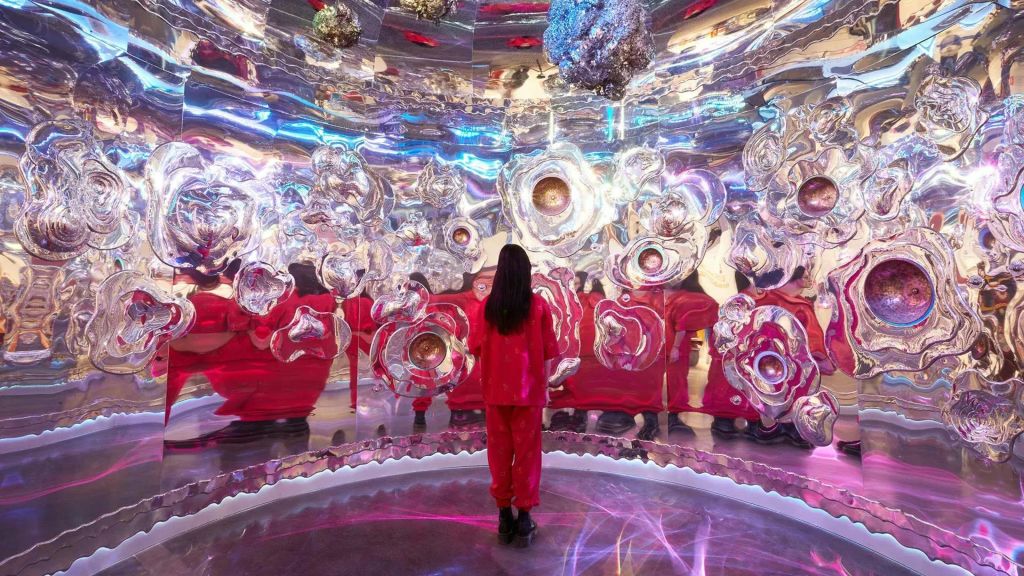 빨간색 의상을 입은 사람은 벽과 천장에 추상적인 금속 모양이 있는 몰입형 반사 예술 작품 내부에 서 있습니다.