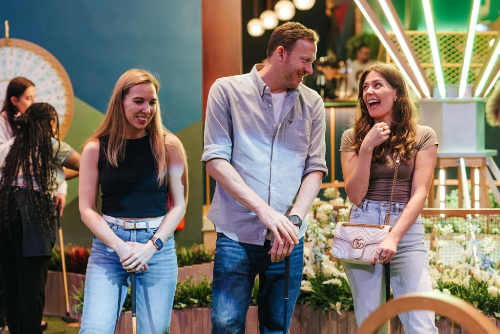 미니 골프 클럽을 든 두 사람이 미소를 짓고 있습니다. 식물과 조명 기구가 보이는 바퀴와 사람들을 배경으로 합니다.