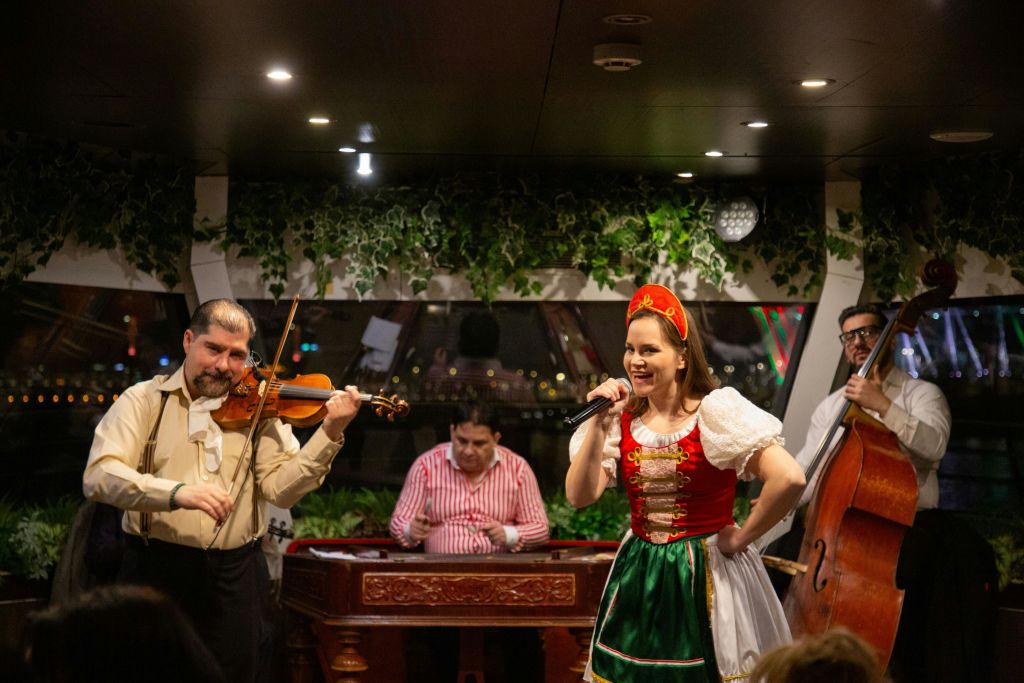 전통 복장을 한 여성이 바이올린, 더블베이스, 해머를 연주하는 라이브 밴드와 함께 장식된 환경에서 노래를 부릅니다.