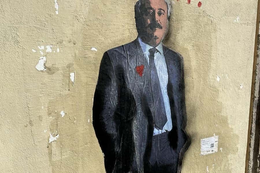 양복을 입은 남성이 주머니에 손을 넣고 질감의 벽을 배경으로 거리 예술을 선보입니다. 빨간색 하트가 옷깃에 있습니다.