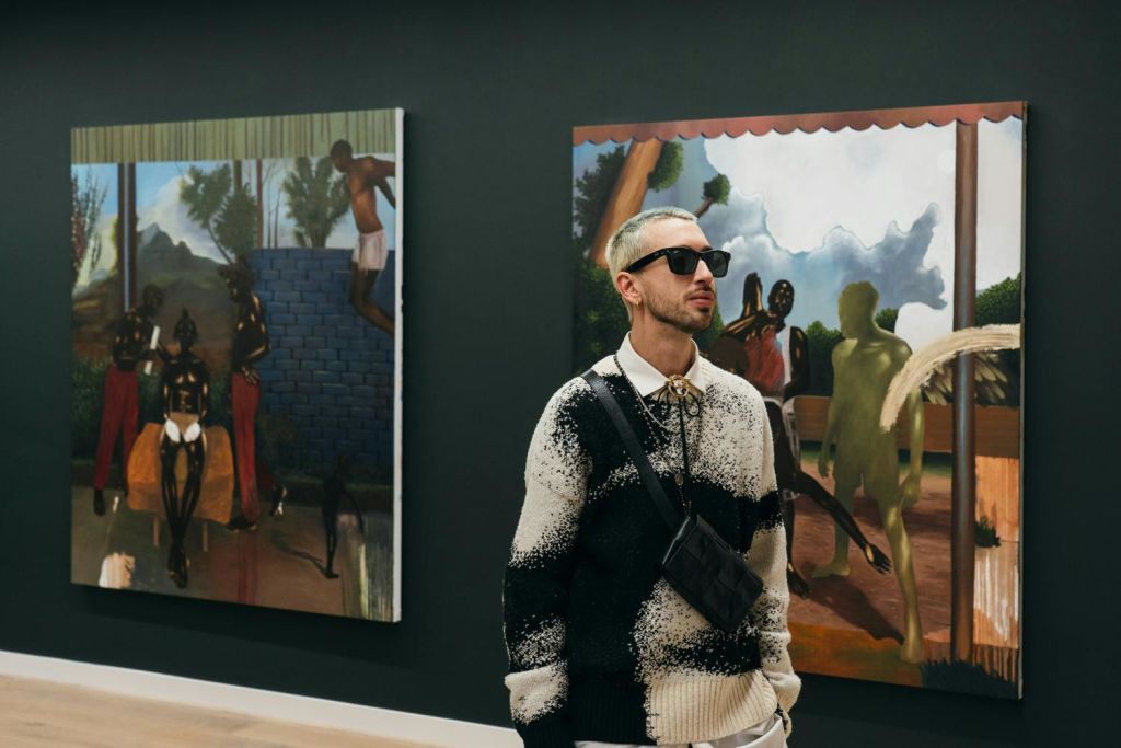 어두운 벽에 걸린 화려한 그림 앞에 선글라스를 쓰고 무늬가 있는 스웨터를 입은 사람이 아트 갤러리에 서 있습니다.