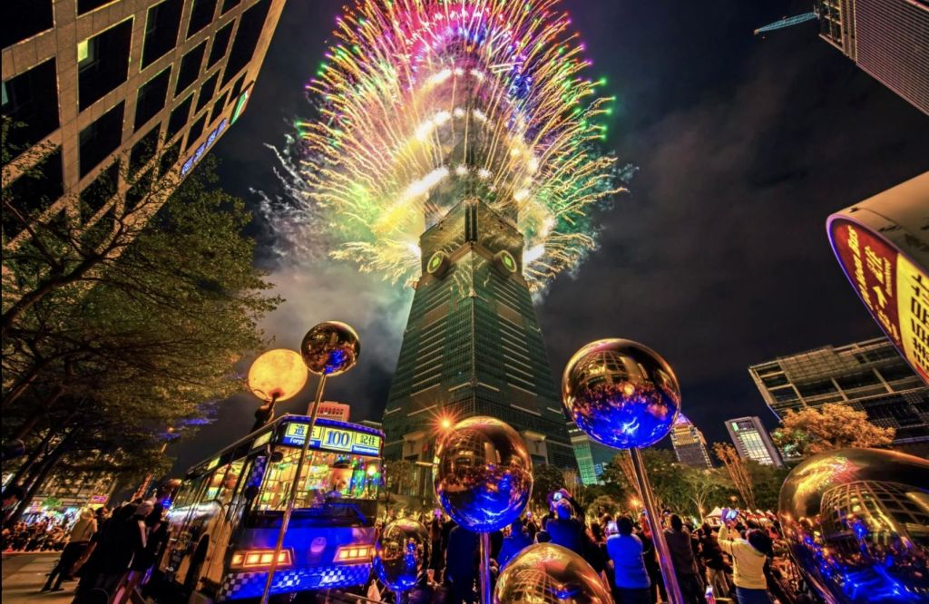 밤에는 높은 건물 위에서 화려한 불꽃놀이가 펼쳐지며, 아래층에는 많은 인파가 몰려들고 전경에는 반사 구체가 펼쳐집니다.