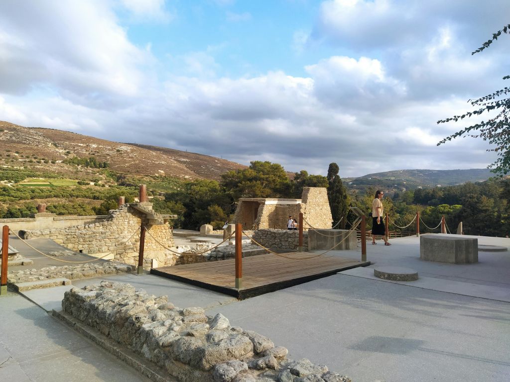 석조 건축물과 목조 산책로, 언덕과 흐린 하늘을 배경으로 걷는 두 사람이 있는 고대 유적지입니다.