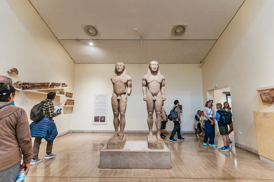 받침대에 서있는 남성 인물의 고대 동상 두 개가 있는 박물관 방. 방문객들은 주변을 관찰하고 돌아다닙니다.