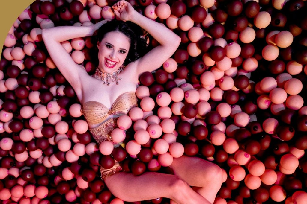 금색 옷을 입은 여성이 분홍색과 갈색 구형 물체가 가득한 수영장에 누워 카메라를 보며 미소를 짓고 있습니다.