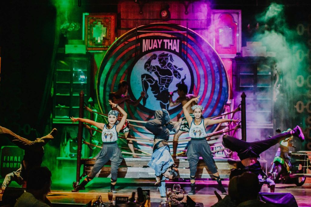 연기 효과가 있는 무대에서 "무에타이" 간판과 화려한 배경이 돋보이는 역동적인 포즈로 춤을 추는 사람들.