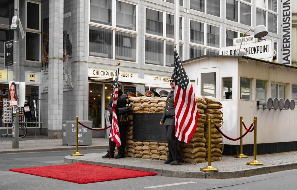 'Checkpoint Charlie' 표지판이 있는 건물 앞에는 두 명의 경비원이 모래주머니와 미국 국기를 들고 검문소에 서 있습니다.