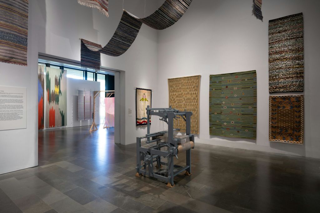 벽에 직조된 직물과 부드러운 조명 아래 전경에 직조 직기가 있는 아트 갤러리입니다.