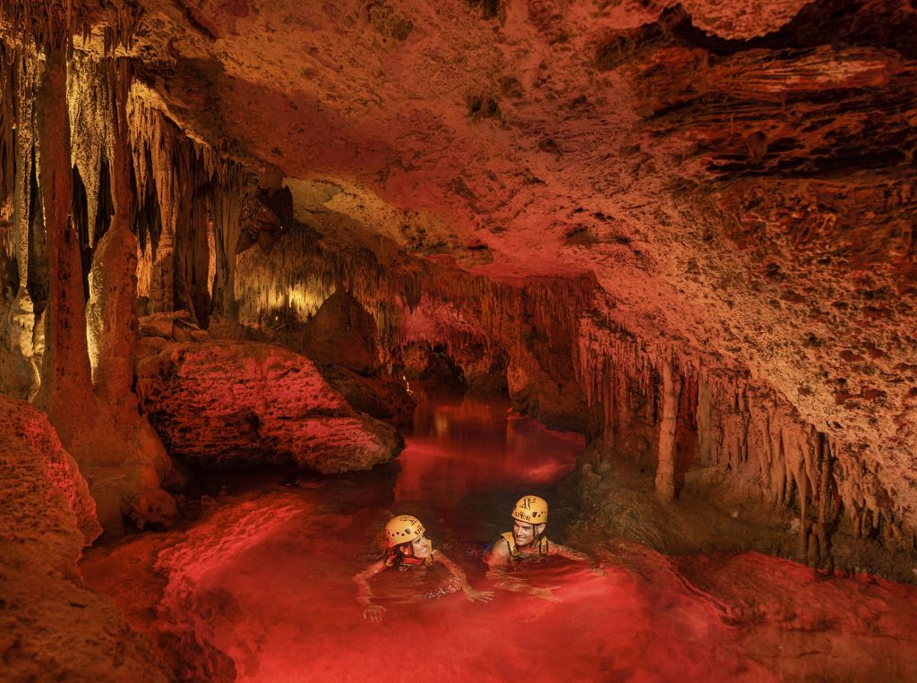 헬멧을 쓴 두 사람이 종유석과 암석으로 둘러싸인 붉은 조명이 있는 물이 가득한 동굴을 지나고 있습니다.