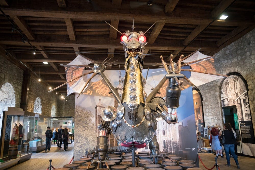 붉게 빛나는 눈과 날개가 달린 금속 용 조각이 실내에 전시되어 있으며, 여러 사람이 배경을 관찰하고 있다.
