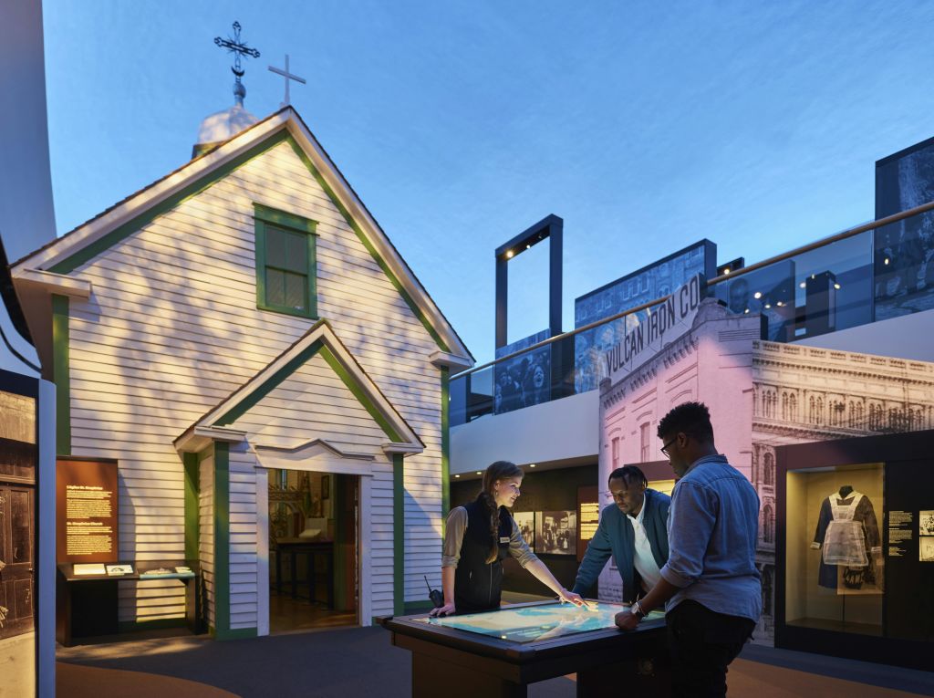 역사적 건물과 유물로 둘러싸인 박물관 전시관에서 세 사람이 조명이 희미한 디스플레이 테이블을 살펴보고 있습니다.