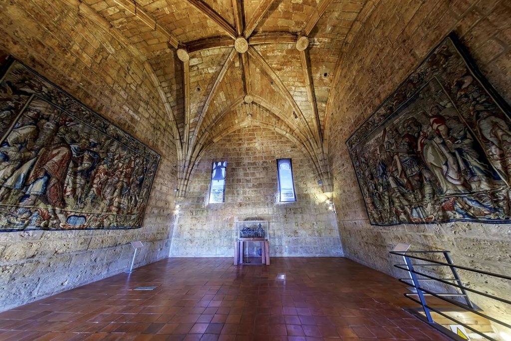 아치형 천장, 창문 2개, 커다란 태피스트리 2개, 타일 바닥, 중앙에 작은 나무 테이블이 있는 돌 벽으로 마감된 객실입니다.