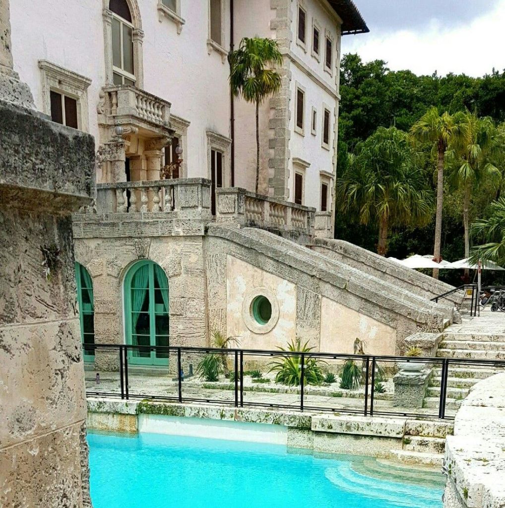 야자수로 둘러싸인 전경에는 아치형 창문, 계단, 청록색 수영장이 있는 유서 깊은 석조 건물이 있습니다.