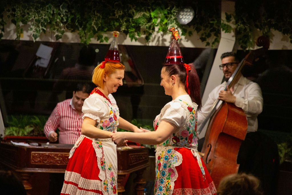 전통 복장을 한 두 여성이 꽃무늬가 있는 빨간색과 흰색 드레스를 입고 파티 모자를 쓰고 춤을 추고 있습니다. 음악가가 배경에서 연주합니다.