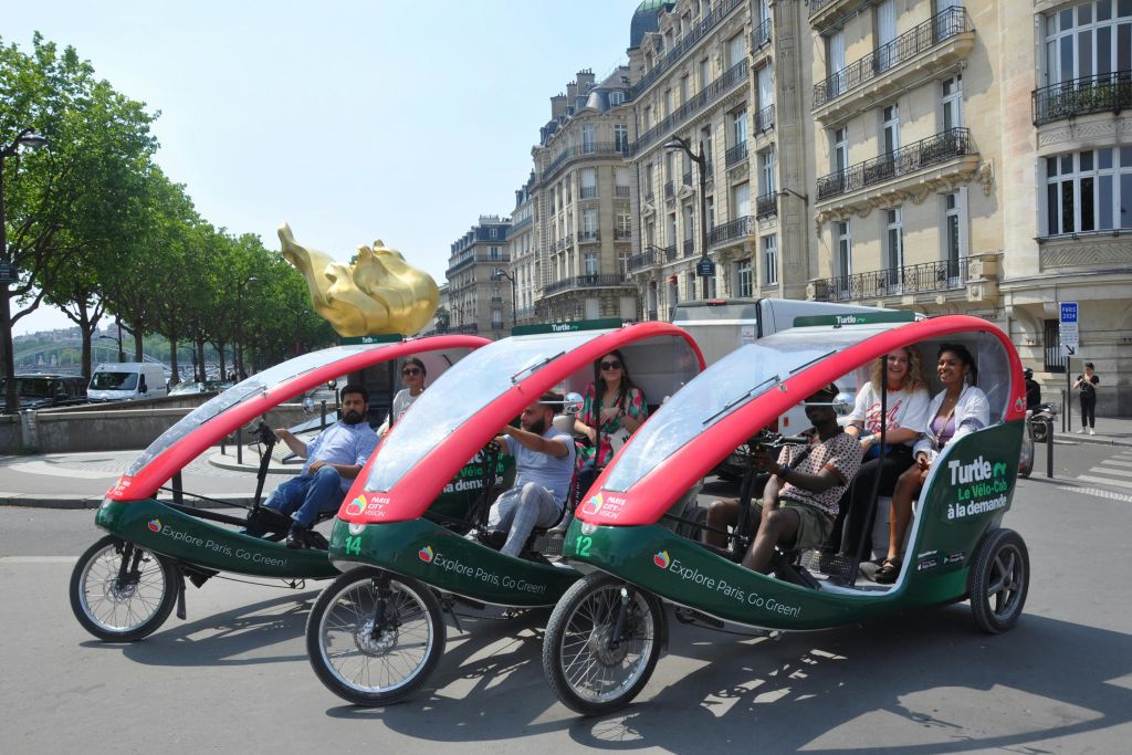 초록색과 빨간색 페디캡 세 대와 역사적인 건물이 있는 파리 거리를 지나가는 승객들.