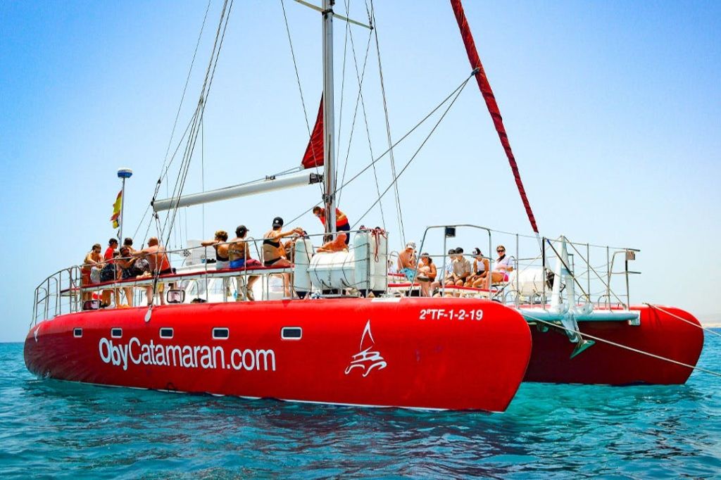 선체에 웹사이트 이름과 범선 로고가 새겨진 푸른 바다의 붉은 쌍동선을 탄 사람들.