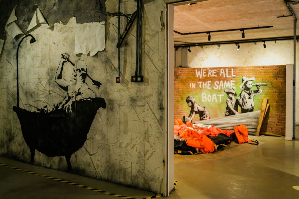 그래피티 벽화와 주황색 구명조끼로 가득한 방이 있는 내부 공간 벽에 "우리는 모두 같은 배에 타고 있습니다" 라는 문구가 적혀 있습니다.