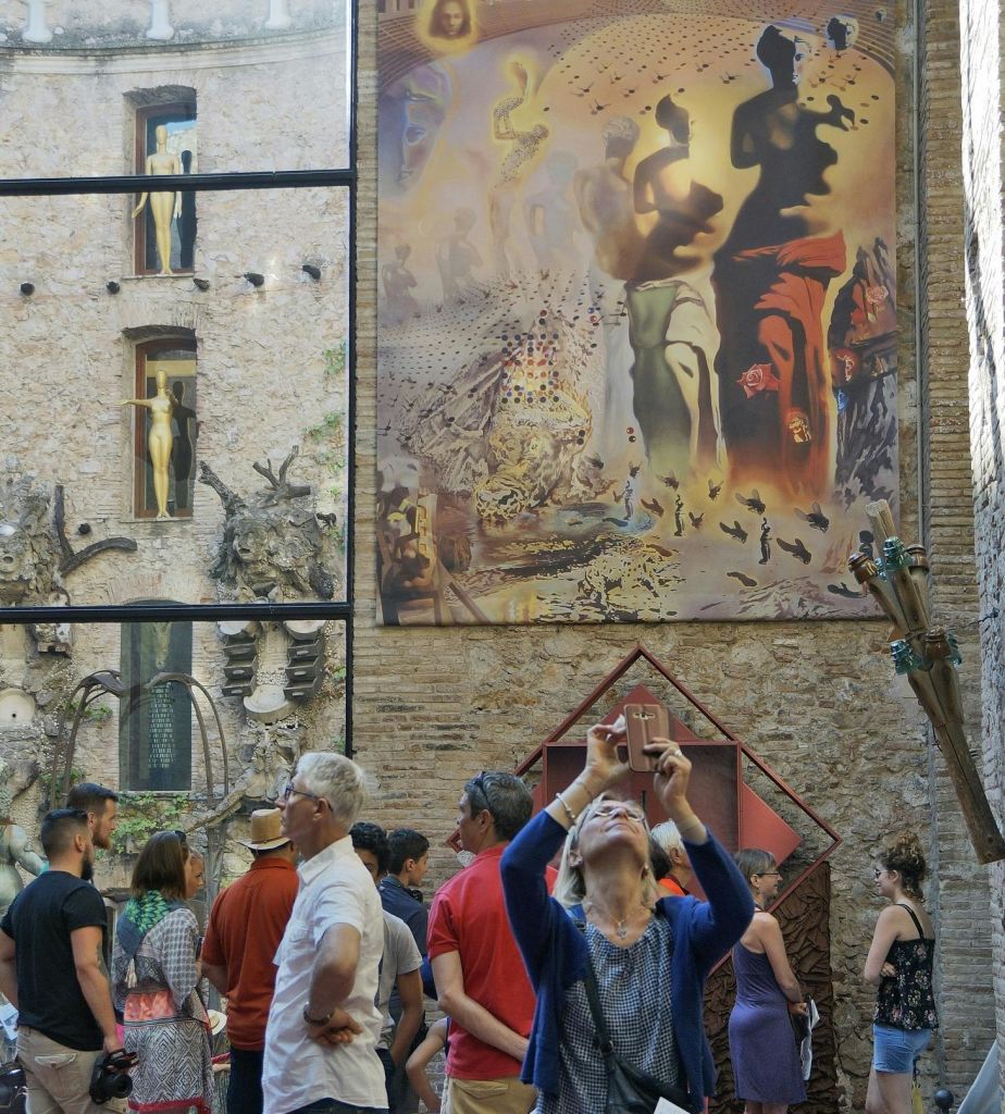 마당에 앉아 있는 한 무리의 사람들, 커다란 벽 벽화를 올려다보는 사람들, 전경에 앉아 있는 한 여성이 사진을 찍고 있는 모습.