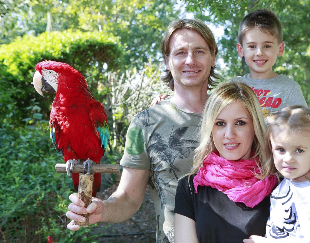 4인 가족이 한 남자의 팔에 붉은 앵무새를 얹은 채 야외에 서 있습니다. 나무와 녹지가 배경입니다.