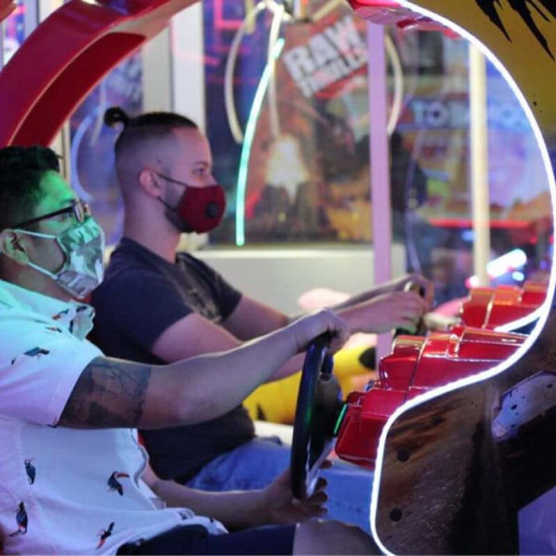 마스크를 쓴 두 사람이 밝고 다채로운 게임 공간에서 레이싱 아케이드 게임을 하고 있습니다.