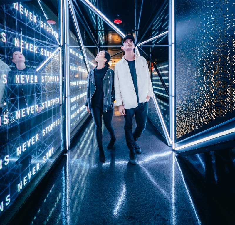 두 사람이 네온 불빛과 거울 벽이 있는 미래형 복도를 거듭 걷고 있습니다.