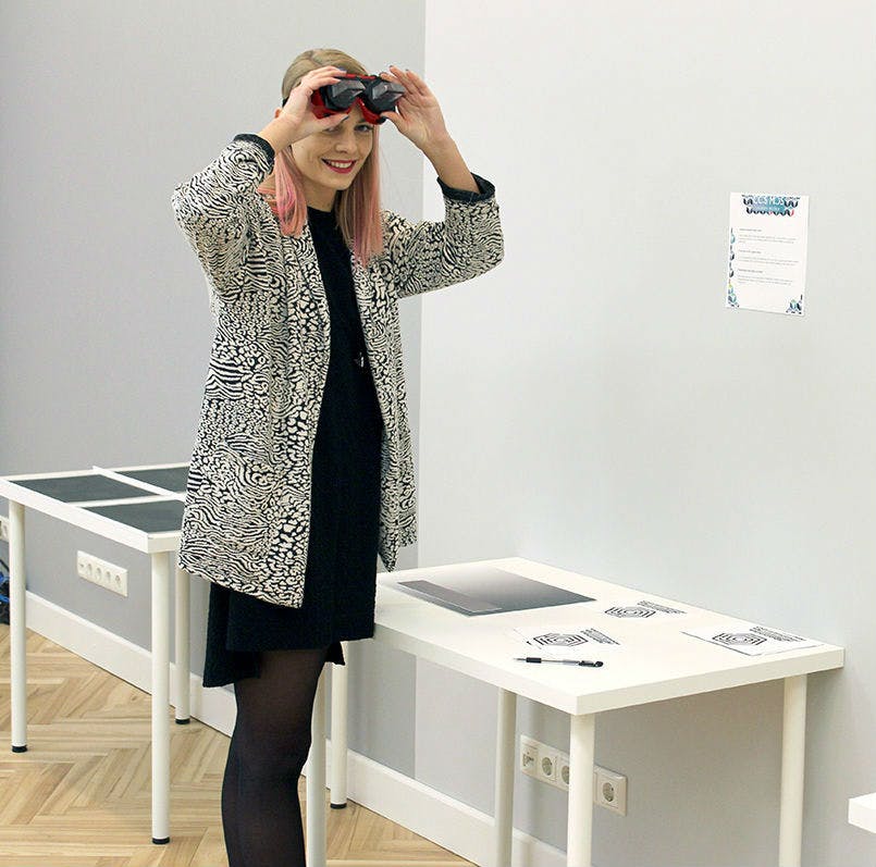 테이블 옆에 서서 무늬가 있는 코트를 입고 미소를 짓고 빨간색과 검은색 가상 현실 헤드셋을 눈에 들고 있는 사람.