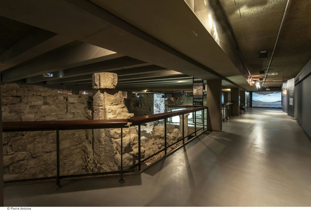 지하 박물관에는 고대 석조 유적, 금속 난간, 정보 패널이 콘크리트 천장 아래에 전시되어 있습니다.