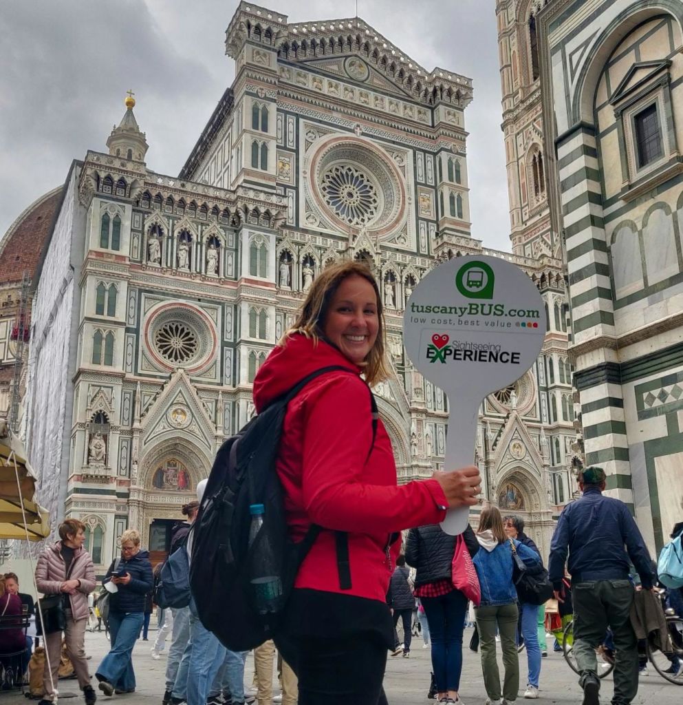 화려한 피렌체 대성당을 배경으로 분주한 광장에서 "tuscanyBUS.com" 간판을 들고 있는 빨간 재킷을 입은 여성.