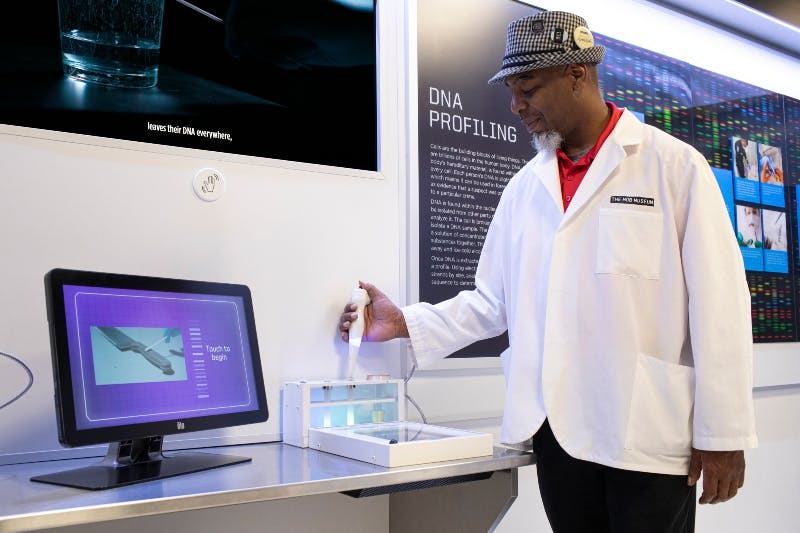 실험복을 입은 사람은 실험실 환경에서 DNA 프로파일링 기계를 작동하며, 벽 뒤에 정보 패널이 있습니다.