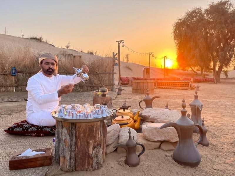 하얀 전통 복장을 한 남성이 해 질 녘 사막의 쿠션에 앉아 나무 테이블 위에 놓인 컵에 냄비의 차를 붓고 있습니다.