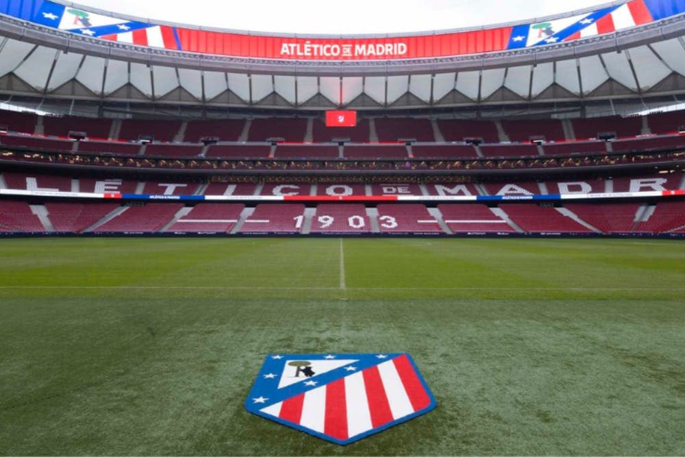 붉은 좌석을 갖춘 축구 경기장, '아틀레티코 데 마드리드' 라고 적힌 커다란 표지판, 잔디에 새겨진 팀 엠블럼.