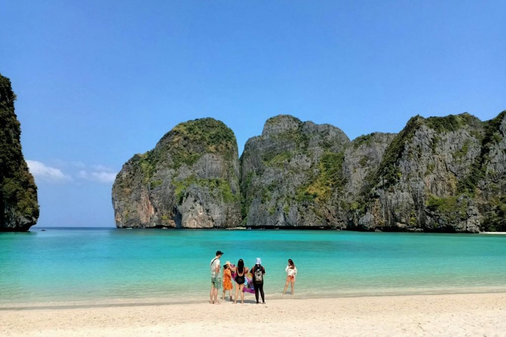 한 무리의 사람들이 청록색 물과 커다란 녹색 절벽을 배경으로 맑고 푸른 하늘 아래 모래 해변에 서 있습니다.
