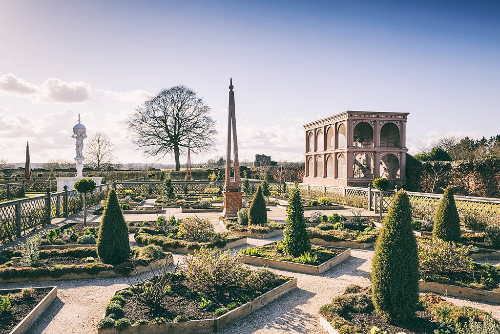 깔끔하게 다듬어진 울타리, 오벨리스크, 맑은 하늘 아래 커다랗고 탁 트인 석조 구조물이 있는 정식 정원입니다.