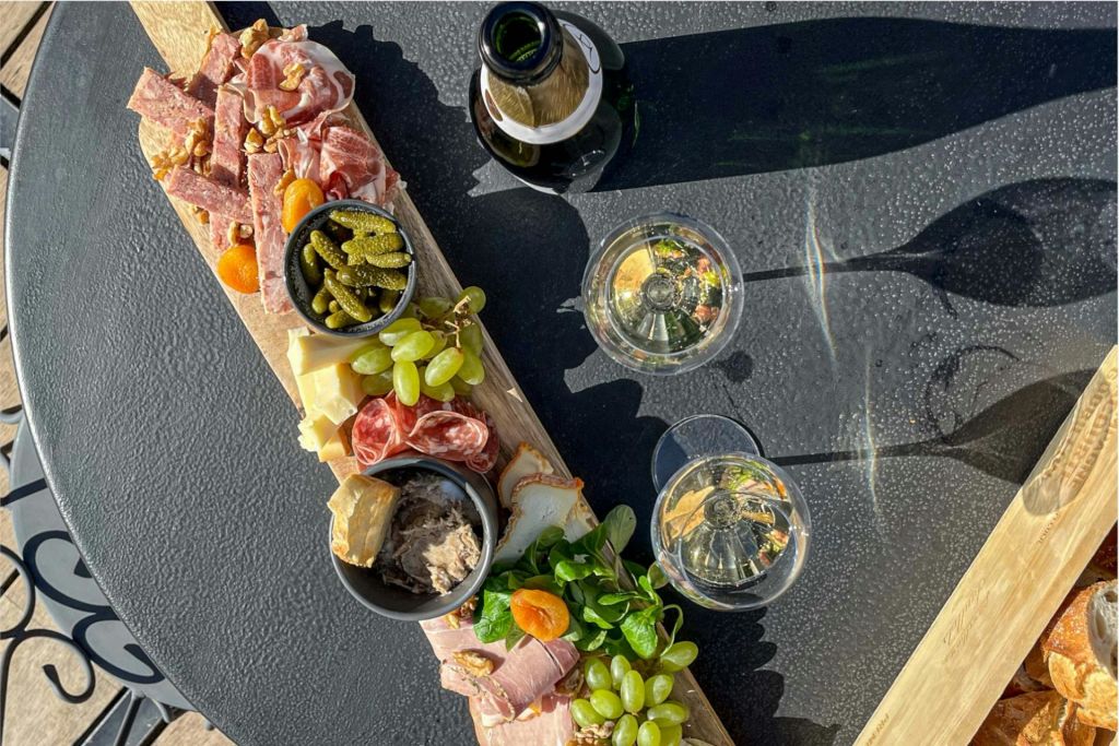 와인병 옆에 고기, 치즈, 포도, 피클이 놓인 샤퀴트리 보드와 검은색 테이블 위에 채워진 잔 두 개가 놓여 있습니다.