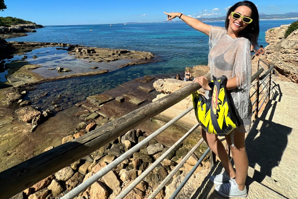 선글라스를 착용한 여성이 화창한 날 난간 너머로 구명조끼를 입은 채 바위 길에 서서 바다를 가리키고 있습니다.