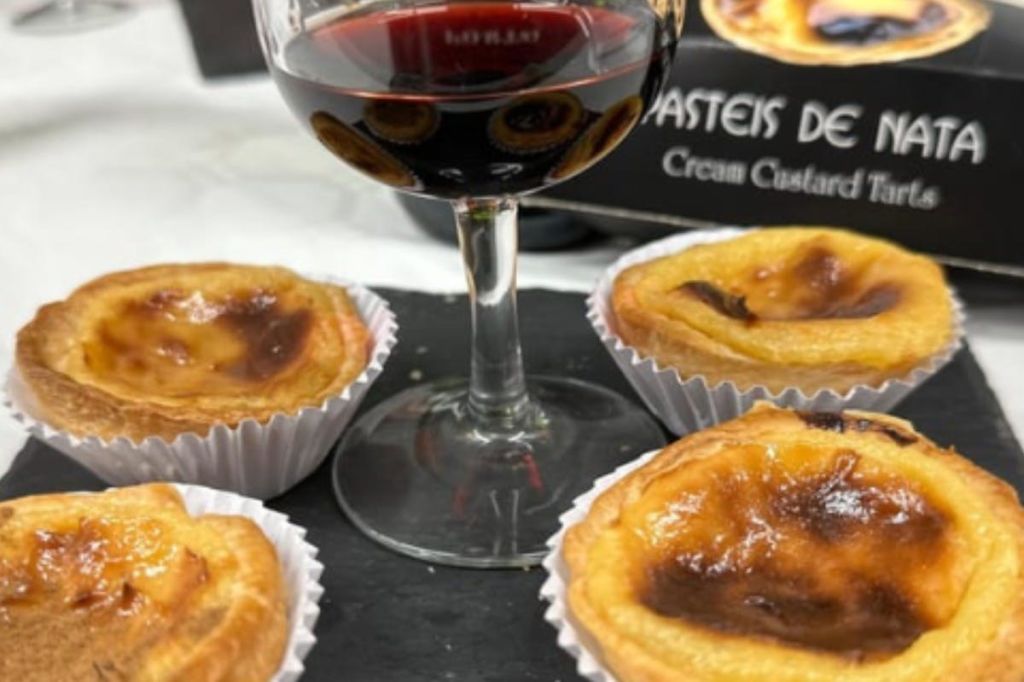 검은색 표면에 레드 와인 한 잔과 포르투갈 커스터드 타르트 네 개가 놓여 있고, 상자 일부가 배경에 보입니다.