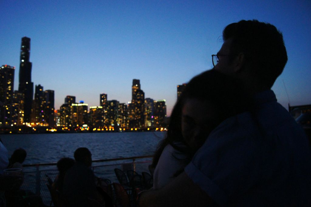 해질 무렵, 물줄기를 가로지르는 도시의 스카이라인을 배경으로 한 커플이 포옹하고 있습니다.