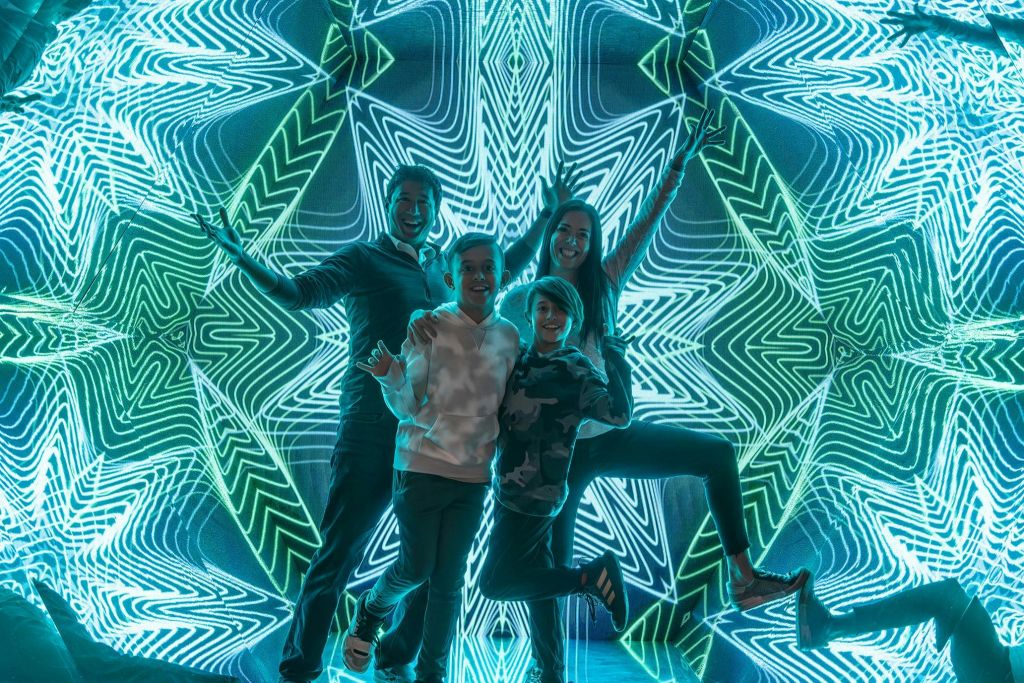 네 사람이 정교하고 대칭적인 네온 패턴으로 빛나는 배경 앞에서 활기차게 포즈를 취하고 있습니다.