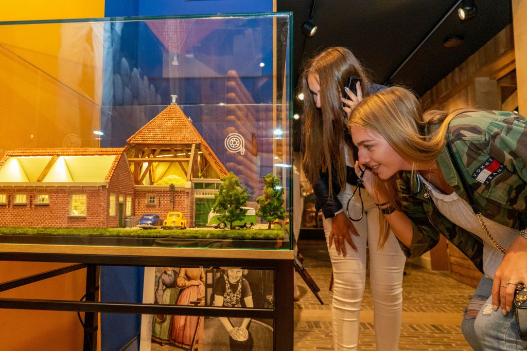두 명의 여성이 건물과 나무의 미니어처 모델을 특징으로 하는 유리 디스플레이 케이스를 살펴보고 오디오 가이드를 듣습니다.
