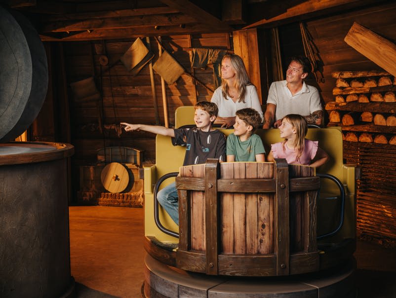 5인 가족이 테마파크 차량에 앉아 웃으며 화면 밖의 무언가를 바라보고 있습니다. 배경은 나무 오두막과 비슷합니다.