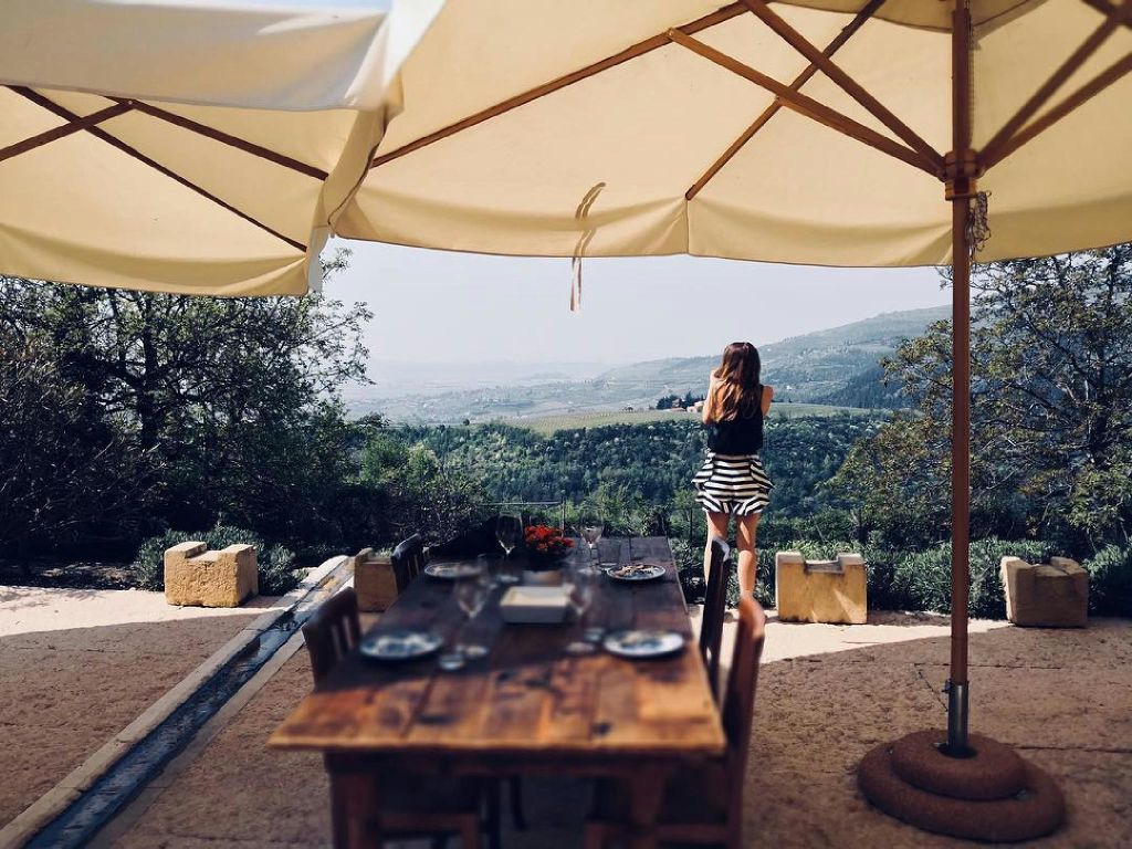한 여성이 커다란 우산 아래 야외 식탁 근처에 서서 아름다운 계곡과 먼 언덕을 내려다보고 있습니다.