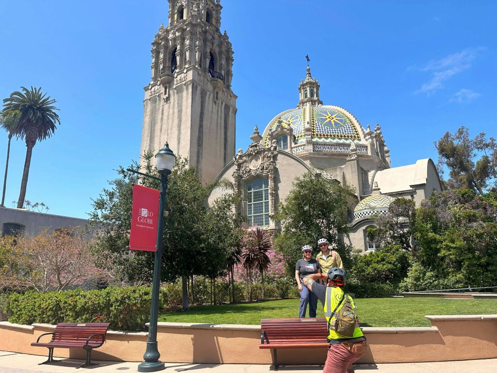 높은 타워와 화려한 돔이 있는 유서 깊은 건물 앞에서 안전 장비를 착용한 사람이 자전거 타는 사람 2명의 사진을 찍고 있습니다.