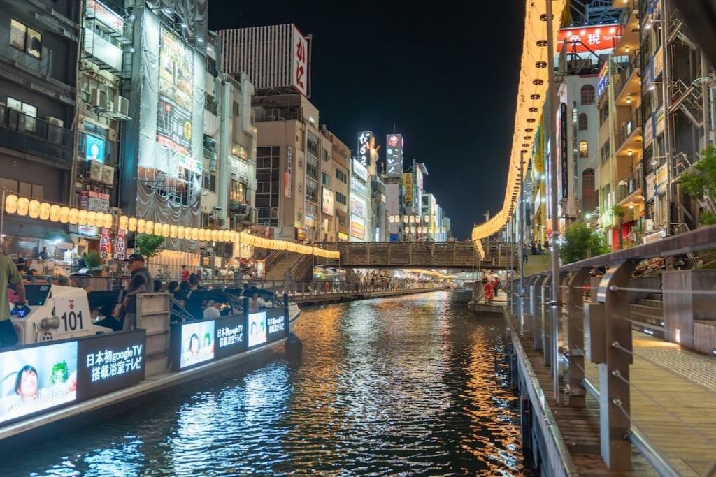 밤에 불이 켜진 건물과 광고판이 늘어서 있는 운하로, 사람들이 물가를 따라 걷고 멀리 다리가 있습니다.