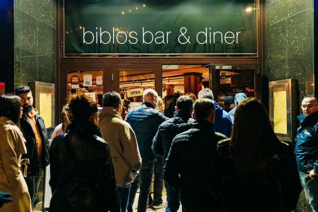 밤에 술집과 식당 밖에서 많은 사람들이 기다리고 있는데, 문에 '열기' 표시가 보입니다.