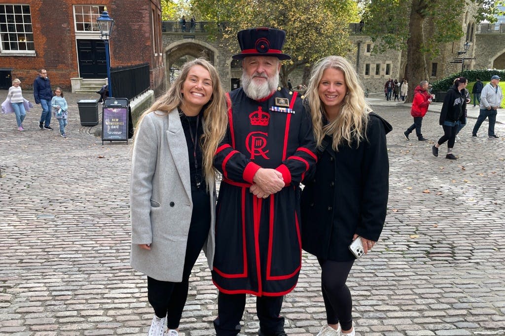 유서 깊은 석조 건물과 나무를 배경으로 자갈길에서 전통 제복을 입은 두 여성이 비피터와 포즈를 취하고 있습니다.