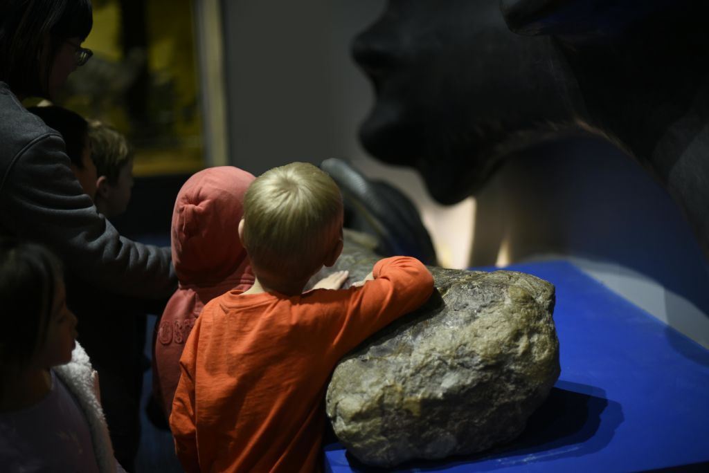 빨간 셔츠를 입은 두 어린이가 박물관의 전광판을 관찰하고 한 어린이가 파란색 표면의 큰 바위를 만지고 있습니다.
