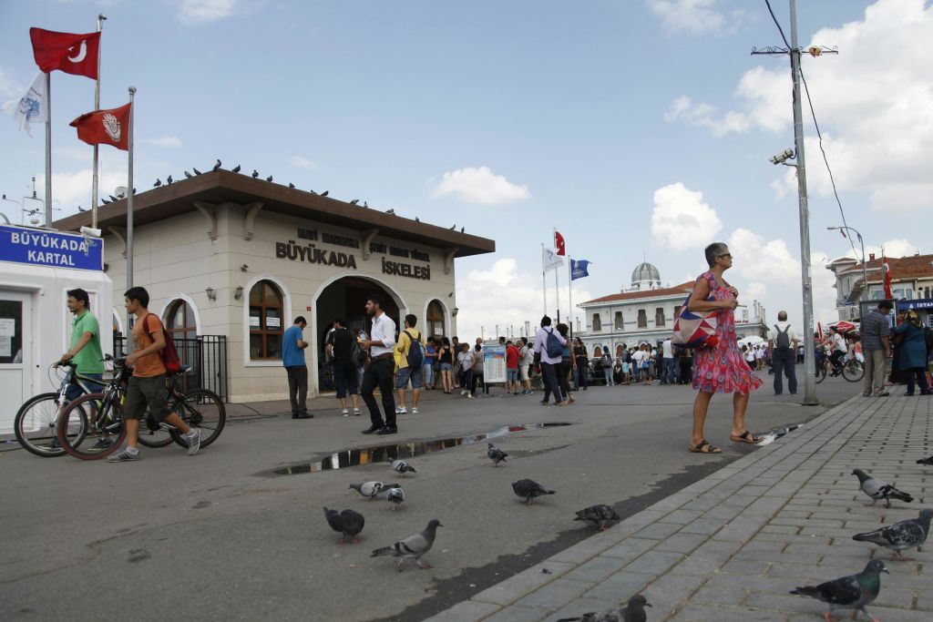 Büyükada İskele 페리 터미널 주변에 사람들이 모여 있습니다. 비둘기가 바닥에 있고 배경에는 깃발이 보입니다.