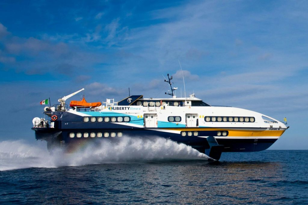 커다란 흰색과 파란색의 리버티 라인 (Liberty Lines) 페리가 물 위를 빠르게 가로지르며 부분적으로 흐린 하늘 아래 스프레이를 만듭니다.