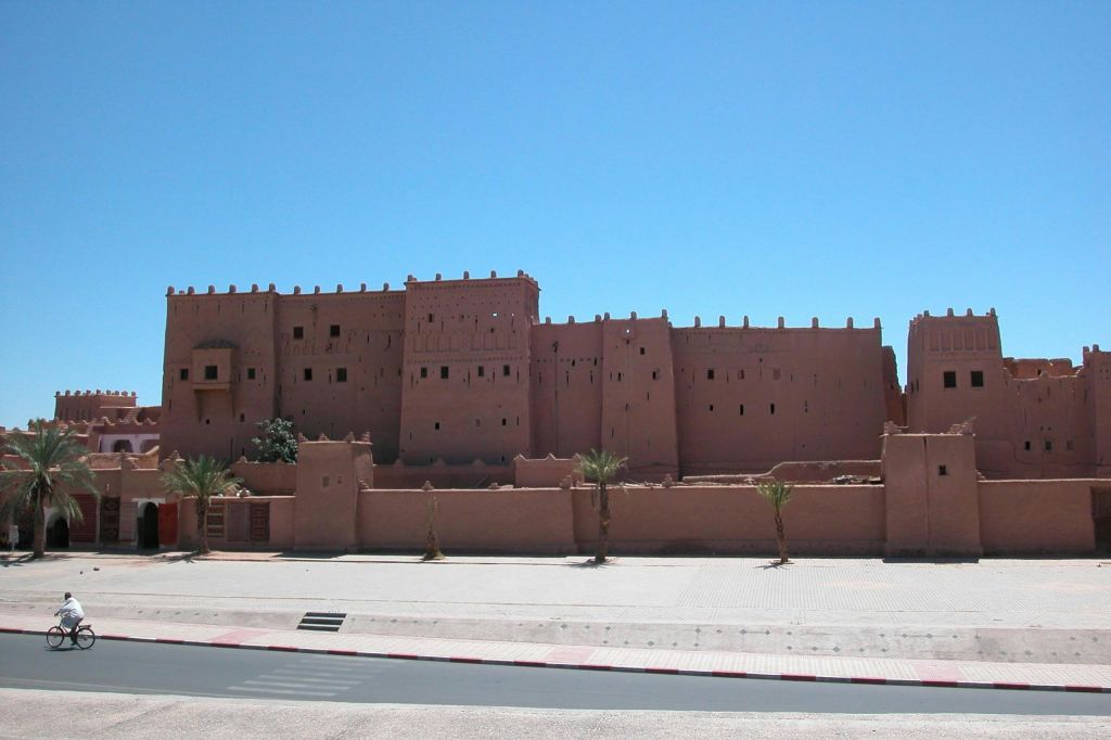 크고 갈색의 진흙 벽돌로 된 요새로, 맑고 푸른 하늘 아래 작은 창문과 야자수가 앞에 있습니다.